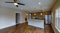 This single-story space showcases an open-concept layout that seamlessly connects the living area and kitchen, creating a spacious and functional environment. The kitchen serves as the focal point, featuring warm medium-toned wood cabinetry that extends to the upper walls, offering generous storage. A large kitchen island with a light granite countertop provides additional workspace and serves as a natural gathering point. Stainless steel appliances, including a range with hood and refrigerator, complement the modern aesthetic. Three pendant lights hang above the island, adding both style and targeted illumination, while recessed lighting throughout ensures the space remains bright and well-lit. Rich, dark hardwood-style flooring runs continuously across the open floor plan, grounding the space with warmth and visual continuity. The living area is equipped with a ceiling fan, contributing to comfort and airflow. Neutral gray walls provide a clean, versatile backdrop throughout. A doorway visible from the main area leads to an adjacent room, suggesting a well-connected and practical floor plan. Overall, the space balances contemporary design elements with a warm, inviting ambiance.