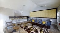 This single-floor space presents a well-designed lounge or common area that combines modern aesthetics with functional versatility. The room is anchored by a striking vaulted ceiling with clean white paneling, which enhances the sense of openness and airiness throughout. Recessed lighting provides even, ambient illumination across the interior.

A bar-height counter runs along one wall, accompanied by a row of sleek adjustable stools in a warm metallic finish, creating an ideal setting for casual work or conversation. Opposite this area, a cluster of modular grey and yellow upholstered chairs arranged around small tables offers a comfortable seating zone, with mobile casters that allow easy reconfiguration.

A large decorative wall panel in a textured golden-yellow hue serves as a bold focal point, complemented by a framed artwork depicting a suspension bridge, adding depth and character to the space. The wood-look flooring grounds the room warmly, while a large statement area rug in dark brown with circular dot patterns in white and yellow introduces a dynamic, contemporary visual element.

The overall ambiance is modern and inviting, with a consistent color palette of grey, yellow, and white that ties the various furnishings and design elements together cohesively, making it a versatile and visually engaging environment.