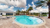 Located at 1517 Cameron Samuel Lane in West Palm Beach, Florida, this outdoor amenity space showcases a beautifully designed resort-style pool area that embodies the relaxed elegance of South Florida living. The centerpiece is a generously sized swimming pool with a graceful curved shape and striking turquoise water, framed by a broad decorative paver deck that invites lounging and leisure. Tall, mature palm trees line the perimeter, providing a tropical ambiance and partial shade throughout the day. Poolside lounge chairs and seating areas are thoughtfully positioned to take full advantage of the sunny Florida climate and open sky views. In the background, white-roofed cabana structures and additional residential buildings can be seen, suggesting a well-appointed community setting. The lush, manicured landscaping and open layout create a welcoming, resort-like atmosphere perfect for relaxation and outdoor recreation. The combination of the vibrant blue pool, sun-drenched pavers, and swaying palms against a backdrop of dramatic Florida clouds makes this space a standout outdoor amenity within the West Palm Beach community.