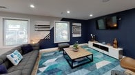 This single-level amenity space is thoughtfully designed to offer a variety of recreational and wellness functions within one cohesive layout. The fitness area is a standout feature, showcasing a bold teal accent wall, multiple treadmills, cable machines, medicine ball storage, and wall-mounted screens, all set atop a striking geometric patterned floor in shades of blue, tan, and gray that flows throughout the space. Recessed lighting illuminates the area evenly, while roller shade windows provide natural light.

Adjacent to the fitness zone, the lounge area features deep navy accent walls contrasted by light wood-look tile flooring. A comfortable sectional sofa, upholstered ottoman, and wood-framed coffee table are anchored by a teal geometric area rug, with a wall-mounted TV and curated abstract artwork completing the look. A kitchenette nearby offers white cabinetry, a teal countertop, under-counter beverage refrigerator, and a wood-plank accent wall. The space also includes a casual dining nook with a round wood table and wire chairs, as well as a game area featuring a pool table. Large windows throughout ensure ample natural light and outdoor views.