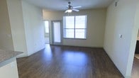 This single-floor residence offers a clean, functional layout designed for comfortable everyday living. The main living area immediately draws attention with its rich, dark hardwood-style flooring that contrasts elegantly against the neutral, light-toned walls. A ceiling fan with an integrated light fixture is centrally positioned overhead, providing both air circulation and ambient lighting throughout the space.

Natural light flows generously into the living area through a large multi-pane window fitted with horizontal blinds, allowing for adjustable light control and privacy. A secondary window to the side further enhances the brightness of the room. A door with its own window panel leads to what appears to be a private balcony or patio, offering a seamless connection between the interior and the outdoors.

The walls are finished in a warm, neutral tone that complements the flooring and contributes to an open, airy atmosphere. Clean baseboards line the perimeter of the room, adding a polished, well-maintained appearance. The open layout of the living space provides generous room to arrange furnishings flexibly. Overall, the space presents a bright, tidy, and inviting environment suited for a range of living arrangements.