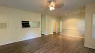 This single-floor space presents an open layout with a cohesive, neutral color palette throughout. The main living area is anchored by rich, dark hardwood-style flooring that contrasts cleanly against light-toned walls, creating a warm yet understated atmosphere. A ceiling fan with a light fixture occupies the center of the room, providing both illumination and air circulation, while a track lighting fixture along the far wall offers additional directed lighting options.

A distinctive built-in nook along one wall houses a microwave, paired with a mirrored backsplash that adds a subtle reflective element to the space. This feature serves as a practical pass-through area, partially separating the living space from adjacent areas. White painted walls are complemented by matching baseboards and door trim, contributing to a clean, uniform finish throughout.

Partial walls and open doorways visible in the background suggest additional rooms beyond the main living area, hinting at a well-connected floor plan. The overall ambiance is bright, neutral, and move-in ready, offering a versatile canvas suitable for a variety of interior styles and furnishing arrangements.