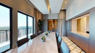 This refined single-floor space is designed with a contemporary aesthetic that balances elegance and functionality. The centerpiece of the space is a generously sized, polished marble dining table surrounded by upholstered charcoal chairs, capable of accommodating a large number of guests. Recessed tray ceilings with embedded lighting add architectural depth and a sense of elevated grandeur to the room.

Floor-to-ceiling windows line one wall, bathing the interior in abundant natural light while offering expansive outdoor views through to an adjoining balcony. Sheer layered curtains frame the windows, providing both privacy and a soft, airy quality to the atmosphere. A tall, lush indoor plant anchors one corner, adding a natural, organic element to the otherwise polished environment.

The space transitions seamlessly into an adjacent kitchen or bar area, visible through a stylish opening framed by patterned drapery panels. This secondary area features warm-toned cabinetry and a striking stone countertop, complemented by curated wall art. The flooring carries a bold geometric pattern that adds visual interest and grounds the overall design. Together, the architectural details, quality materials, and thoughtful layout create a space that is both functional and impressively appointed.