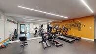 This fitness studio is designed to energize and motivate, offering a comprehensive workout environment across a single open floor. The space is anchored by a bold, amber-orange accent wall emblazoned with fitness-themed typography — featuring words like "Strength," "Endurance," "Warrior," and "Relentless" — creating a high-energy focal point that sets the tone for the entire room. A secondary white wall carries the phrase "Will It Be," reinforcing the motivational aesthetic throughout.

The flooring consists of durable, textured rubber tiles in a dark gray tone, providing cushioning and traction suitable for a wide range of exercises. Overhead, sleek linear LED lighting runs the length of the ceiling, delivering bright, uniform illumination across the workout area. Full-length mirrors along one wall visually expand the space and allow users to monitor their form during training.

The cardio section is well-appointed with multiple treadmills, elliptical machines, and stationary bikes, including a dedicated spin cycle setup. A Bowflex multi-functional strength machine anchors a separate strength training alcove, complemented by a dumbbell rack and additional functional training equipment. Natural light filters in through windows near the entrance, adding to the open, welcoming atmosphere of this fully outfitted fitness facility.