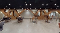 This expansive single-floor lounge makes an immediate architectural statement through a series of large, riveted steel truss structures that traverse the open space in an X-pattern formation. Finished in a warm, oxidized copper-brown tone, these industrial-inspired installations serve as the defining design element, dividing the floor plan into distinct functional zones while maintaining an open, flowing atmosphere.

The polished concrete flooring runs continuously throughout, complementing the dark ceiling panels punctuated by recessed lighting and pendant fixtures. In the main seating area, a cluster of oversized globe pendant lights hangs at varying heights above an eclectic mix of leather and upholstered armchairs in amber, charcoal, and neutral tones, creating an inviting gathering space. Bistro-height tables paired with bentwood stools occupy zones between the trusses, ideal for casual conversation or collaborative work.

A dedicated kitchenette features warm wood cabinetry with dark countertops, an undermount sink, and pendant lighting overhead, accented by a dramatic curtain of string lights cascading from an adjacent truss. A quieter alcove offers a wall-mounted television alongside colorful contemporary chairs in cream, yellow, and magenta for a more intimate retreat. Throughout, the combination of industrial structure and curated furnishings creates a dynamic, design-forward environment.
