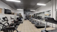 This fitness center at 250 W 5th St in Cincinnati, OH, offers a well-equipped, single-floor workout space designed for cardiovascular training. The room features an open layout with a plaid-patterned resilient floor, providing a clean and functional environment for exercise. Overhead fluorescent lighting illuminates the space evenly, complemented by an exposed ceiling with visible ductwork that adds an industrial aesthetic.

The cardio equipment selection is comprehensive, including multiple treadmills, elliptical trainers, upright stationary bikes, and stair climbers, all arranged in organized rows to maximize usable floor space. Full-length mirrors line the walls throughout the room, visually expanding the space while allowing users to monitor their form during workouts. A wall-mounted flat-screen television is positioned for easy viewing during exercise sessions.

The facility includes gray storage lockers along one wall, providing secure storage options for users. A water dispenser and waste receptacle are conveniently located near the entrance, and a sanitizing wipe dispenser is mounted on the wall for equipment hygiene. The entryway features a glass-panel door with an automatic closer, ensuring smooth access. The overall design is practical and modern, prioritizing functionality and efficient use of space.