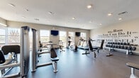 Located at 383-389 Main St, this single-floor fitness facility offers a thoughtfully designed layout comprising an exercise room, a living room, and a connecting hallway. The generously sized exercise room serves as the centerpiece of the space, featuring durable rubber flooring, recessed lighting, and large windows that invite abundant natural light while offering outdoor views. The room is outfitted with a comprehensive selection of cardio and strength-training equipment, including treadmills, ellipticals, stationary bikes, cable machines, free weights, and adjustable benches. Floor-to-ceiling mirrors along one wall enhance the sense of openness and allow users to monitor their form. Wall-mounted flat-screen TVs provide entertainment during workouts, and a motivational typography mural adds an energetic, encouraging atmosphere. The living room area provides a welcoming lounge space with warm hardwood-style flooring, a boldly patterned geometric accent wall, and a wall-mounted display screen — ideal for relaxation or group viewing. The hallway connects the two areas seamlessly, completing a functional and modern fitness environment designed to support an active lifestyle.