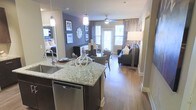 This single-floor residence offers an open-concept layout that seamlessly connects the kitchen, dining, and living areas, creating a cohesive and functional living environment. The kitchen serves as a central anchor of the space, featuring a large granite-topped island with an undermount sink, dark cabinetry, stainless steel appliances including a dishwasher, and elegant pendant lighting that adds a modern, polished touch. Hardwood-style flooring flows continuously throughout, lending warmth and visual continuity across all areas. The dining zone is positioned just off the kitchen, providing a natural transition into the living area, which is generously sized and bathed in natural light from large windows and a glass door. The living room is finished with neutral tones and accented by a ceiling fan, a floor lamp, and thoughtfully arranged seating that reinforces the space's welcoming ambiance. An accent wall in a deep gray-blue hue adds depth and character to the interior. Decorative framed artwork and a sunburst mirror enhance the walls, contributing to the space's curated aesthetic. A glimpse of a bedroom beyond the living area suggests well-proportioned private quarters that complement the open communal spaces.