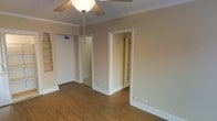 This single-floor space presents a clean, functional layout with warm hardwood-style flooring running throughout. The main room is finished with neutral beige walls, white crown molding, and white baseboards, lending a classic and tidy aesthetic. A ceiling fan with a light fixture is centrally mounted, providing both airflow and ambient lighting to the space.

Along one wall, built-in shelving units offer convenient open storage, ideal for books, decor, or organizational purposes. Two doorways lead from the main room — one opening to what appears to be a hallway or adjoining room, and the other revealing a spacious walk-in closet fitted with multiple built-in shelves, providing ample storage capacity.

The overall ambiance is modest and move-in ready, with a straightforward design that prioritizes practicality. The neutral color palette and consistent flooring throughout create a cohesive look, while the crown molding adds a subtle architectural detail that elevates the space slightly above a purely utilitarian feel. The layout is well-suited for versatile use, whether as a bedroom or a multipurpose room.