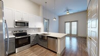 This well-designed 2-bedroom, 2-bathroom residence offers a thoughtful layout across a single floor, blending modern finishes with functional living spaces. The open-concept kitchen serves as the heart of the home, featuring a large center island with a white quartz countertop, stainless steel farmhouse sink, and a full suite of stainless steel appliances including a gas range, built-in microwave, dishwasher, and French door refrigerator. Two-tone cabinetry — white upper cabinets paired with gray lowers — is complemented by a mosaic tile backsplash and pendant lighting that hangs from impressively high ceilings. The kitchen flows seamlessly into the living area, which is brightened by natural light from sliding glass doors and a ceiling fan overhead. Wood-style flooring runs throughout the entire residence, adding warmth and continuity. Both bedrooms offer a clean, neutral palette with natural light from slatted blinds. The primary bedroom connects to an en-suite bathroom with a dual-sink vanity featuring gray cabinetry and white countertops, a glass-enclosed walk-in shower with tile surround, and direct access to a spacious walk-in closet outfitted with built-in shelving and dual hanging rods. A second full bathroom and additional closet provide convenience for guests or a second occupant.