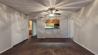 This single-floor residence offers a well-organized layout with distinct living, kitchen, sleeping, and bathroom areas. The living room serves as the heart of the home, showcasing rich dark hardwood-style flooring, a textured ceiling, a ceiling fan with integrated lighting, and a classic fireplace as a focal point. Vertical blinds along a large window allow for adjustable natural light. A pass-through window connects the living area to the kitchen, maintaining an open and connected feel. The galley-style kitchen is efficiently designed with warm wood cabinetry lining both walls, light-toned countertops, a double sink, and black appliances including a gas range, microwave, dishwasher, and refrigerator — providing ample storage and workspace. The bedroom features plush carpeting, a ceiling fan, a window with blinds that brings in natural light, and neutral walls that offer a calm, restful atmosphere. An additional carpeted room with a window provides flexible use as a second bedroom or office. The bathroom includes a wood-toned storage cabinet and continues the dark hardwood-style flooring found throughout the main living spaces. Throughout the residence, neutral tones and consistent finishes create a cohesive and functional living environment.