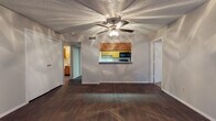 This single-floor residence offers a well-organized layout with distinct living, kitchen, sleeping, and bathroom areas. The living room serves as the heart of the home, showcasing rich dark hardwood-style flooring, a textured ceiling, a ceiling fan with integrated lighting, and a classic fireplace as a focal point. Vertical blinds along a large window allow for adjustable natural light. A pass-through window connects the living area to the kitchen, maintaining an open and connected feel. The galley-style kitchen is efficiently designed with warm wood cabinetry lining both walls, light-toned countertops, a double sink, and black appliances including a gas range, microwave, dishwasher, and refrigerator — providing ample storage and workspace. The bedroom features plush carpeting, a ceiling fan, a window with blinds that brings in natural light, and neutral walls that offer a calm, restful atmosphere. An additional carpeted room with a window provides flexible use as a second bedroom or office. The bathroom includes a wood-toned storage cabinet and continues the dark hardwood-style flooring found throughout the main living spaces. Throughout the residence, neutral tones and consistent finishes create a cohesive and functional living environment.
