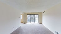 This single-floor space presents a clean, understated interior centered around a spacious main living area. The room is finished with wall-to-wall carpeting in a neutral tone, contributing to a quiet and comfortable atmosphere. A textured popcorn ceiling adds subtle depth overhead, while the crisp white walls provide a bright, versatile backdrop throughout.

The most prominent feature of the living area is a wide sliding glass door that spans much of the exterior wall, offering direct access to an outdoor balcony or patio. This generous glazing draws in abundant natural light and frames a lush, tree-lined view, creating a strong visual connection to the outdoors. A wall-mounted HVAC vent is positioned nearby, ensuring efficient climate control throughout the space.

An adjacent room, visible through an open doorway, benefits from its own window, further enhancing the natural light available across the unit. The overall layout appears open and flowing, with a straightforward floor plan that maximizes usable space. The neutral color palette and practical finishes throughout create a functional, move-in ready environment suitable for a variety of uses.