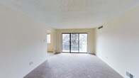 This single-floor space presents a clean, understated interior centered around a spacious main living area. The room is finished with wall-to-wall carpeting in a neutral tone, contributing to a quiet and comfortable atmosphere. A textured popcorn ceiling adds subtle depth overhead, while the crisp white walls provide a bright, versatile backdrop throughout.

The most prominent feature of the living area is a wide sliding glass door that spans much of the exterior wall, offering direct access to an outdoor balcony or patio. This generous glazing draws in abundant natural light and frames a lush, tree-lined view, creating a strong visual connection to the outdoors. A wall-mounted HVAC vent is positioned nearby, ensuring efficient climate control throughout the space.

An adjacent room, visible through an open doorway, benefits from its own window, further enhancing the natural light available across the unit. The overall layout appears open and flowing, with a straightforward floor plan that maximizes usable space. The neutral color palette and practical finishes throughout create a functional, move-in ready environment suitable for a variety of uses.