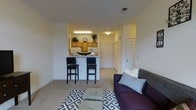 This single-floor unit offers an open and functional layout that seamlessly connects the living area to the kitchen. The living space is carpeted and comfortably sized, providing room for a sofa and seating arrangement centered around a patterned area rug. Recessed into the layout, the kitchen features warm wood-toned cabinetry, white appliances including a refrigerator and microwave, and a peninsula-style breakfast bar with two bar stools — ideal for casual dining or entertaining. The kitchen is well-lit with overhead fluorescent lighting, complementing the warmer ambient lighting throughout the unit. White walls and standard-height ceilings contribute to a clean, neutral aesthetic throughout. A hallway visible from the living area leads to additional rooms, with white panel doors lining the corridor. The entryway is clearly defined, offering a practical transition from the main living spaces into private areas of the unit. Overall, the space presents a straightforward and efficient floor plan with a cohesive, understated design well-suited for everyday living.