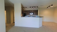 This single-floor residence showcases a thoughtfully designed open-concept layout that connects the kitchen and living areas in a seamless, functional flow. The kitchen is a standout feature, outfitted with rich dark cabinetry that provides generous storage space, black appliances including a refrigerator and built-in microwave, and a dark countertop that adds a sleek contrast to the neutral surroundings. A peninsula-style breakfast bar extends into the living space, offering a casual dining or seating area and serving as a natural dividing element between the kitchen and the main living zone. Track lighting runs along the kitchen ceiling, delivering focused illumination that enhances both the functionality and the ambiance of the cooking area. The living space is finished with plush wall-to-wall carpeting in a warm neutral tone, contributing to a comfortable and inviting atmosphere. Light, neutral-colored walls throughout keep the space feeling open and airy. A hallway visible in the background hints at additional rooms beyond the main living area. Overall, the space presents a clean, modern aesthetic with a practical layout well-suited for everyday living.