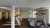 This fitness center is a dedicated, single-floor exercise facility designed to meet a range of workout needs. The space features a practical and clean aesthetic, with a drop-panel ceiling equipped with recessed lighting that provides even, consistent illumination throughout. The flooring is finished in dark rubber matting, a durable and slip-resistant surface well-suited for high-traffic gym use.

The cardio section is prominently featured and includes multiple treadmills and elliptical machines, offering ample options for aerobic training. A multi-station cable machine anchors the strength training area, providing versatility for a wide range of resistance exercises. The equipment is arranged to allow comfortable movement between stations.

A wall-mounted split air conditioning unit ensures climate control, keeping the environment comfortable during intense workouts. Structural support columns are integrated into the layout without significantly impeding the usable floor space. Natural light filters in through a window on one side of the room, complementing the artificial lighting and contributing to an open, energizing atmosphere.

Overall, this fitness center is a functional and well-maintained space that provides users with the essential tools needed for both cardiovascular and strength-based training.