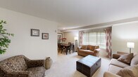 This thoughtfully designed one-bedroom, one-bathroom residence offers a comfortable and efficient single-floor layout. The open living and dining area is bright and welcoming, featuring plush wall-to-wall carpeting, neutral warm-toned walls, and large windows dressed with horizontal blinds and decorative patterned curtains that allow abundant natural light to fill the space. The living room comfortably accommodates multiple seating arrangements and flows seamlessly into the dining area, which provides ample space for a full dining table and chairs. The galley-style kitchen is well-equipped with warm wood cabinetry, dark granite-style countertops, a stainless steel sink, black appliances including a refrigerator and range, and tile flooring — offering excellent functionality within a compact footprint. The bedroom is a serene retreat with soft carpeting, neutral walls, and enough room for a full furniture suite including a dresser and nightstands. The bathroom features classic white subway tile surrounding a full tub-shower combination, a wood-toned vanity with a white countertop, and tasteful accent tile details. A dedicated closet provides additional storage. Overall, the space presents a clean, comfortable, and practical layout suited for everyday living.