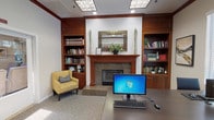 This single-floor space blends a professional office environment with the warm, inviting character of a traditional library. The main office area is anchored by a large dark-wood desk equipped with dual monitors, complemented by ergonomic seating and a printer workstation positioned beneath a window overlooking lush greenery. Natural light filters through slatted blinds, adding brightness to the neutral-toned carpeted floors and warm beige walls accented by rich dark crown molding.

A standout feature of the space is the elegant wood-framed fireplace with a granite surround, flanked symmetrically by rich cherry-wood built-in bookshelves stocked with an extensive collection of books and decorative accents. A decorative mosaic mirror is mounted above the fireplace mantel, which is adorned with potted greenery and framed art.

An adjacent lounge area, accessible through a set of glass-paneled double doors, provides comfortable yellow patterned accent chairs and additional built-in shelving, creating a welcoming reception or waiting space. A second dedicated workstation area features a wall-length white built-in bookcase filled with a diverse collection of books, offering ample reference resources. Recessed and panel lighting throughout ensures a well-lit, functional environment suitable for both focused work and collaborative use.