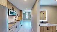 This thoughtfully designed single-floor unit blends modern aesthetics with functional living. The open-plan layout connects the kitchen and living area seamlessly, creating a bright, airy atmosphere enhanced by light hardwood-style flooring that runs throughout the main space. Large windows frame striking urban skyline views, filling the living area with natural light while roller shades offer privacy when needed. A distinctive reclaimed wood accent column adds a touch of industrial character to the living space.

The kitchen is efficiently laid out along one wall and is outfitted with wood-toned cabinetry featuring matte black hardware, smooth quartz-style countertops, a stainless steel undermount sink with a sleek black faucet, a built-in cooktop, stainless steel appliances including an over-the-range microwave, a dishwasher, and a white subway tile backsplash that adds clean contrast to the warm wood tones.

The bathroom is finished with large-format neutral floor tiles and features a glass-enclosed walk-in shower with tile surround, a modern vanity with a matte black faucet, and matte black accessory hardware throughout, creating a cohesive, contemporary feel. The overall palette of warm neutrals, natural wood textures, and matte black accents gives this unit a polished, modern character.