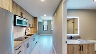 This thoughtfully designed single-floor unit blends modern aesthetics with functional living. The open-plan layout connects the kitchen and living area seamlessly, creating a bright, airy atmosphere enhanced by light hardwood-style flooring that runs throughout the main space. Large windows frame striking urban skyline views, filling the living area with natural light while roller shades offer privacy when needed. A distinctive reclaimed wood accent column adds a touch of industrial character to the living space.

The kitchen is efficiently laid out along one wall and is outfitted with wood-toned cabinetry featuring matte black hardware, smooth quartz-style countertops, a stainless steel undermount sink with a sleek black faucet, a built-in cooktop, stainless steel appliances including an over-the-range microwave, a dishwasher, and a white subway tile backsplash that adds clean contrast to the warm wood tones.

The bathroom is finished with large-format neutral floor tiles and features a glass-enclosed walk-in shower with tile surround, a modern vanity with a matte black faucet, and matte black accessory hardware throughout, creating a cohesive, contemporary feel. The overall palette of warm neutrals, natural wood textures, and matte black accents gives this unit a polished, modern character.