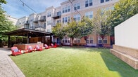 Located in Alexandria, Virginia, this inviting outdoor courtyard serves as a central gathering space within a modern multi-story residential community. The courtyard is defined by a well-maintained artificial turf lawn that provides a clean, green focal point throughout the seasons. Mature trees are thoughtfully planted throughout the space, offering natural shade and a sense of established greenery that softens the surrounding architecture.

A standout feature of the courtyard is the covered outdoor kitchen and lounge area, constructed beneath a sleek contemporary pergola with a louvered metal roof. The outdoor kitchen is anchored by a brick-faced bar counter, equipped with grilling stations and ample prep space, making it ideal for community gatherings and social events. String lights are draped overhead, adding a warm, ambient atmosphere for evening use.

Surrounding the outdoor kitchen, vibrant red Adirondack chairs with cushions are arranged to encourage relaxed conversation and casual lounging. A large projection screen is mounted on one end of the courtyard, suggesting the space is used for outdoor movie screenings and community events.

Herringbone-patterned brick walkways connect various areas of the courtyard, complemented by neatly landscaped planting beds featuring ornamental grasses and shrubs. The surrounding multi-story buildings feature balconies and large windows that overlook the courtyard, creating an integrated indoor-outdoor living experience.