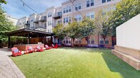 Located in Alexandria, Virginia, this inviting outdoor courtyard serves as a central gathering space within a modern multi-story residential community. The courtyard is defined by a well-maintained artificial turf lawn that provides a clean, green focal point throughout the seasons. Mature trees are thoughtfully planted throughout the space, offering natural shade and a sense of established greenery that softens the surrounding architecture.

A standout feature of the courtyard is the covered outdoor kitchen and lounge area, constructed beneath a sleek contemporary pergola with a louvered metal roof. The outdoor kitchen is anchored by a brick-faced bar counter, equipped with grilling stations and ample prep space, making it ideal for community gatherings and social events. String lights are draped overhead, adding a warm, ambient atmosphere for evening use.

Surrounding the outdoor kitchen, vibrant red Adirondack chairs with cushions are arranged to encourage relaxed conversation and casual lounging. A large projection screen is mounted on one end of the courtyard, suggesting the space is used for outdoor movie screenings and community events.

Herringbone-patterned brick walkways connect various areas of the courtyard, complemented by neatly landscaped planting beds featuring ornamental grasses and shrubs. The surrounding multi-story buildings feature balconies and large windows that overlook the courtyard, creating an integrated indoor-outdoor living experience.