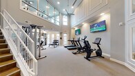 This fitness center is defined by its dramatic double-height ceilings, which lend the space an open, airy character reinforced by recessed lighting and ceiling fans that maintain a comfortable environment throughout. A mezzanine-level glass railing overlooks the main workout floor below, adding architectural interest while maximizing the sense of vertical space.

The main floor is organized into distinct functional zones. The cardio area is equipped with multiple treadmills, an elliptical trainer, a stationary bike, and a rowing machine, all arranged to face wall-mounted flat-screen televisions. The strength training zone features a multi-station cable machine by Hoist, a selection of free weights on tiered racks, and an adjustable workout bench, supplemented by large mirrored walls that visually expand the space and aid in form correction.

Natural light enters through several tall, arched windows and a set of French doors that open to the exterior, contributing to a bright and welcoming atmosphere. The neutral-toned carpeted flooring and soft gray walls create a clean, modern aesthetic throughout. A separate vestibule area with a staircase and glass-panel door provides access to additional levels, completing this comprehensive and thoughtfully designed fitness facility.