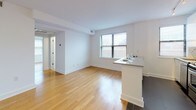 This well-designed single-floor residence offers a clean, contemporary aesthetic throughout, characterized by crisp white walls, warm hardwood flooring, and a thoughtfully organized layout. The open-concept kitchen is a standout feature, outfitted with sleek white cabinetry, stainless steel appliances including a gas range and microwave, a subway tile backsplash, and a peninsula with a quartz countertop that doubles as a breakfast bar. Dark tile flooring anchors the kitchen zone, creating a clear visual distinction from the adjoining living area. The living space is generously proportioned, bathed in natural light from multiple windows fitted with horizontal blinds. Its clean lines and open floor plan provide a versatile canvas for a variety of furniture arrangements. A convenient in-unit stacked washer and dryer is neatly tucked behind cabinetry, adding a practical touch. The bedroom features warm hardwood floors and a wide closet with a full-length hanging rod, offering ample storage. The bathroom is finished with large-format neutral stone-toned tiles, a soaking tub, built-in corner shelving, and a safety grab bar, combining function with refined detail. Throughout the unit, the consistent use of white tones and natural wood creates a bright, cohesive, and welcoming atmosphere.