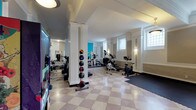 This fitness center combines classic architectural character with a fully equipped workout environment. Distinctive white fluted columns rise from a polished checkerboard terrazzo floor, lending the space an elegant, timeless aesthetic that sets it apart from a typical gym. The open layout allows for an unobstructed flow between workout zones, accommodating a variety of exercise equipment including stationary bikes, an elliptical trainer, a rowing machine, and a multi-station strength machine. A medicine ball rack stationed between the columns offers a range of weighted balls for functional training exercises. Large frosted windows along one wall allow natural light to filter into the space while maintaining privacy, and wall-mounted sconces provide supplemental illumination. A full-length mirror along one side of the room enhances the sense of depth and allows for form-checking during workouts. A "Healthier 4U" healthy vending machine near the entrance offers convenient access to nutritious snacks and beverages. The walls feature a crisp white finish accented by turquoise panels, adding a vibrant, energetic touch to the overall atmosphere. The space is cooled by a wall-mounted air conditioning unit, ensuring comfort during intense workouts.