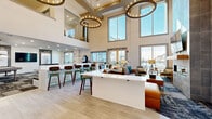 This contemporary two-story amenity clubhouse is designed to impress from the moment you enter. The main level is anchored by a breathtaking double-height great room, where soaring ceilings and an expansive wall of large windows flood the space with natural light and offer views of the surrounding community. Dramatic circular chandelier fixtures suspended at varying heights add warmth and visual interest overhead. A sleek white bar-height island serves as the centerpiece, complemented by stylish wood and metal bar stools, while a full-height tile-clad fireplace wall with built-in shelving and a mounted television creates a striking focal point in the lounge area. Comfortable seating arrangements encourage relaxed gathering, and light-toned tile flooring flows seamlessly throughout. A dedicated kitchenette area features white cabinetry, quartz countertops, and stainless steel appliances, ideal for entertaining. A game room offers additional recreational space, decorated with curated artwork and access to the outdoors. A separate conference or meeting room provides a quieter setting, appointed with a large table, upholstered bench seating, and refined wall sconce lighting. A modern open-riser staircase with black metal railings connects the two floors, completing the building's polished, community-focused design.
