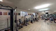 This single-floor fitness center offers a comprehensive workout environment designed to accommodate a variety of exercise routines. The space is finished with durable wood-look flooring throughout, textured ceilings, and a combination of recessed and overhead fluorescent lighting that keeps the area well-lit. A distinctive stacked stone accent wall runs along one side of the room, adding a stylistic element to the otherwise functional space.

The strength training area is well-appointed with a cable cross machine and multiple Life Fitness selectorized machines, including leg and upper-body stations arranged along the stone wall. Natural light filters in through several windows positioned along the walls. The cardio section features multiple treadmills and elliptical trainers lined up along a mirrored wall, which visually expands the space and allows users to monitor their form. A wall-mounted television is positioned for viewing during cardio sessions.

Additional amenities include a stability ball, a collection of free weights, and a ceiling fan for air circulation. Convenient refreshment options are readily available, with a Dasani refrigerated beverage cooler stocked with drinks and a snack vending machine located near the entrance. A wall clock and posted facility guidelines complete the practical touches throughout.