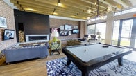 This single-floor amenity lounge is a thoughtfully designed social space that seamlessly blends modern sophistication with warm, inviting textures. The room is anchored by a sleek, linear gas fireplace framed in dark brick, which adds a striking focal point to one wall. Opposite, a full exposed brick wall serves as a gallery backdrop for multiple mounted flat-screen televisions, providing entertainment throughout the space.

A regulation-size billiard table, dressed in a light felt top, takes center stage atop a bold, patterned area rug, making it an immediate draw for guests. Comfortable lounge seating, including plush sofas and accent chairs, is arranged throughout to encourage conversation and relaxation.

Overhead, slatted wood ceiling beams run the length of the room, adding architectural warmth and visual depth, while a contemporary globe chandelier introduces a touch of refined elegance. Large floor-to-ceiling windows flood the interior with abundant natural light and offer open views of the surrounding greenery, creating a seamless connection between indoors and outdoors.

Warm wood-tone flooring ties all the design elements together, lending the space a cohesive, upscale feel. Overall, this lounge is a well-appointed, multi-functional gathering space ideal for socializing and leisure.