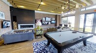 This single-floor amenity lounge is a thoughtfully designed social space that seamlessly blends modern sophistication with warm, inviting textures. The room is anchored by a sleek, linear gas fireplace framed in dark brick, which adds a striking focal point to one wall. Opposite, a full exposed brick wall serves as a gallery backdrop for multiple mounted flat-screen televisions, providing entertainment throughout the space.

A regulation-size billiard table, dressed in a light felt top, takes center stage atop a bold, patterned area rug, making it an immediate draw for guests. Comfortable lounge seating, including plush sofas and accent chairs, is arranged throughout to encourage conversation and relaxation.

Overhead, slatted wood ceiling beams run the length of the room, adding architectural warmth and visual depth, while a contemporary globe chandelier introduces a touch of refined elegance. Large floor-to-ceiling windows flood the interior with abundant natural light and offer open views of the surrounding greenery, creating a seamless connection between indoors and outdoors.

Warm wood-tone flooring ties all the design elements together, lending the space a cohesive, upscale feel. Overall, this lounge is a well-appointed, multi-functional gathering space ideal for socializing and leisure.