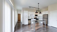 Highland Station presents a thoughtfully designed single-floor residence that combines modern finishes with comfortable, functional living. The open-concept kitchen is a standout feature, showcasing white shaker cabinetry, dark granite countertops, a classic white subway tile backsplash, and a full suite of stainless steel appliances including a gas range, built-in microwave, dishwasher, and refrigerator. Industrial-style pendant lighting adds character above the central island workspace. The kitchen flows seamlessly into a generously sized living area filled with natural light from large windows and a set of French doors. A clean-lined fireplace anchors the living space, adding warmth and elegance. Wide-plank hardwood-style flooring runs throughout the main living areas, while plush carpet provides comfort in the bedrooms. The primary bedroom features a striking black barn door that opens to a spa-like en-suite bath complete with a double-sink vanity, dark granite counters, and a floor-to-ceiling marble-tiled walk-in shower. Spacious walk-in closets with wire shelving systems offer excellent storage. A secondary bathroom mirrors the refined design with white cabinetry and granite countertops. The overall ambiance is bright, contemporary, and move-in ready.