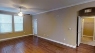 This single-floor space presents a well-maintained interior highlighted by rich, dark hardwood flooring that anchors the room with warmth and character. The walls are finished in a neutral warm beige tone, providing a versatile backdrop adaptable to a variety of furnishing styles. Elegant crown molding runs along the ceiling perimeter, adding a refined architectural detail that elevates the overall aesthetic.

The main living area is generously proportioned, offering ample room for flexible furniture arrangements. A ceiling fan is centrally positioned, contributing to both comfort and air circulation throughout the space. Natural light enters through windows fitted with white horizontal blinds, allowing for adjustable brightness and privacy as needed.

Clean white baseboards provide crisp contrast against both the flooring and walls, contributing to a polished, cohesive look. A door with a window panel, also dressed with blinds, leads to an exterior area, further enhancing the sense of connectivity to the outdoors. The space also features a wall-mounted thermostat, underscoring its practical, move-in-ready condition. Overall, the space exudes a simple, functional charm suited to a range of lifestyles.