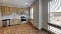 Located at 3705 SW Durham Dr in Durham, NC, this single-floor unit offers a thoughtfully designed, modern living space with a clean, contemporary aesthetic. The open-concept layout connects the living area to the kitchen, creating a seamless flow throughout the main living spaces. Warm wood-look flooring runs consistently across the unit, complementing the neutral gray and white color palette on the walls and trim.

The kitchen is well-appointed with natural wood-toned cabinetry, light quartz countertops, a subway tile backsplash, and a full suite of stainless steel appliances, including a refrigerator with ice maker, electric range, over-the-range microwave, and dishwasher. A window above the counter area allows natural light to fill the space while providing outdoor views.

The living area benefits from large, wide windows fitted with horizontal blinds, inviting generous natural light and views of the surrounding neighborhood. Recessed lighting and ceiling fans provide additional comfort and functionality.

The bathroom is finished with matching wood-toned cabinetry, a white undermount sink, a large mirror with a dark frame, and a walk-in glass-enclosed shower with white tile surround. A hallway with panel-style doors and dark hardware connects the private areas of the unit, maintaining a cohesive and polished look throughout.