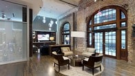 This distinguished space masterfully combines historic architectural elements with refined modern design. The interior is anchored by dramatic exposed brick walls and sweeping arched openings that speak to the building's storied past, while soaring ceilings create an expansive, airy atmosphere throughout. Tall, multi-pane arched windows and full-length glass door panels flood the space with abundant natural light, offering views to the surrounding urban streetscape. The lounge area is thoughtfully appointed with a curated arrangement of contemporary upholstered armchairs in neutral tones, centered around a sleek round glass coffee table and grounded by a plush area rug — all set atop rich hardwood flooring. A reception or media station is positioned toward the rear, complete with a flat-screen display, ambient table lamps, and built-in shelving. Overhead, a cascading cluster of globe pendant lights adds a sculptural, artistic focal point that complements the industrial aesthetic. The interplay of raw brick textures, warm wood tones, polished metal accents, and soft upholstery creates a welcoming yet sophisticated ambiance — ideal for gathering, reception, or communal use.