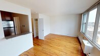 This single-floor space offers a clean, open layout designed for comfortable everyday living. The main living area is highlighted by warm, honey-toned hardwood floors that run throughout, lending the space a timeless and inviting character. Crisp white walls reflect natural light, creating a bright and airy atmosphere that enhances the sense of openness.

A built-in shelving unit provides practical storage and display space, seamlessly integrated into the architecture alongside a classic six-panel interior door. A partial wall with a granite-topped counter visually separates the kitchen area from the living space, offering a functional pass-through design that maintains an open feel while defining distinct zones.

The kitchen area, partially visible from the main living space, features cabinetry and the granite countertop detail, adding a touch of contrast to the otherwise neutral palette. The layout flows naturally, making the space feel connected and easy to navigate.

Overall, the space is characterized by its simplicity, clean finishes, and warm hardwood flooring, creating a versatile and livable environment that is both practical and welcoming. The neutral color scheme and open configuration provide an excellent canvas for a variety of interior styles and furnishing arrangements.