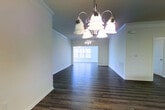 This single-story residence showcases an open-concept layout that seamlessly connects the living area, dining space, and kitchen into one cohesive environment. Dark hardwood-style flooring runs throughout, providing a warm, grounding contrast to the crisp white walls and trim. The living area is anchored by a ceiling fan with an integrated light fixture, ensuring both comfort and illumination across the generous open floor plan.

A pendant chandelier marks the dining area, offering a subtle decorative focal point that distinguishes the space without disrupting the open flow. The kitchen is positioned at the rear, featuring dark wood cabinetry, black appliances including a built-in microwave and range, and a raised breakfast bar that serves as a natural divider between the kitchen and the main living areas. Crown molding detail adds a refined architectural touch along the upper walls, lending a polished character to the interior. The layout is practical and functional, with a clean, neutral palette that provides a versatile backdrop adaptable to a variety of interior design preferences.