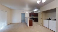 This single-floor space offers an open-concept layout that seamlessly connects the kitchen and living area, creating a functional and cohesive environment. The kitchen is a standout feature, equipped with rich dark wood cabinetry that provides ample storage, complemented by stainless steel appliances including a gas range and built-in microwave. A peninsula with a dark granite-style countertop extends into the living space, offering additional prep area and casual seating potential. A pendant light hangs above the peninsula, adding focused illumination, while a chandelier anchors the adjacent living area. The kitchen flooring transitions to a neutral tile, while the living area is finished with plush, light-toned carpeting that adds warmth underfoot. Recessed and flush-mounted ceiling lighting throughout ensures well-distributed light across the space. A doorway leads to an additional room, suggesting a bedroom or utility area, and a separate door near the kitchen provides access to what appears to be a laundry area, with a white appliance visible. The overall palette of cream walls and neutral tones creates a clean, inviting atmosphere.