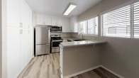 Located at 9601 El Rey Ave in Fountain Valley, California, this single-floor residence offers a clean, modern interior with thoughtful updates throughout. The open living area features light-toned luxury vinyl plank flooring, a textured ceiling, and a ceiling fan, with a sliding glass door that fills the space with natural light and opens to an outdoor area. The layout flows seamlessly into a well-appointed kitchen equipped with stainless steel appliances — including a refrigerator, range, and microwave — complemented by white shaker cabinetry, granite-style countertops, and a stainless steel sink beneath a window with horizontal blinds. A peninsula provides additional counter space and a natural divide between the kitchen and living areas. The bedroom is carpeted and features a window with views of surrounding greenery, offering a quiet and comfortable retreat. The bathroom is finished with matching granite-style countertops, white cabinetry, a well-lit vanity mirror, and durable wood-look flooring. A separate water closet and a shower-tub combination add to the bathroom's functionality. Neutral warm-gray tones are carried throughout the space, creating a cohesive and inviting ambiance well-suited to Fountain Valley's Southern California setting.