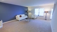This two-floor space showcases a thoughtfully designed interior that balances contemporary style with functional comfort. The main living area immediately draws attention with its striking deep blue accent wall, which creates a bold focal point against the surrounding neutral-toned walls. Soft, wall-to-wall carpeting in a warm beige hue extends throughout the room, adding a sense of comfort underfoot while complementing the overall color palette. Natural light flows generously through large windows, brightening the space and offering views of the surrounding exterior. The living area is well-proportioned, providing ample room for seating arrangements and open movement. The clean lines and minimalist aesthetic are reinforced by white furniture, including a sofa and armchair, paired with a sleek, modern coffee table featuring a mirrored surface. A tall floor lamp with a tripod base adds both height and ambient lighting to the room. The neutral ceiling and trim provide a crisp finish that ties the design elements together cohesively. Spanning two floors, this space offers versatile living arrangements suitable for a variety of lifestyles and needs.