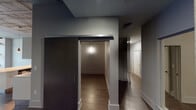 This contemporary single-floor loft showcases an industrial-modern aesthetic defined by striking exposed concrete ceilings, rich hardwood flooring, and an open-concept layout that maximizes the sense of space. The living area is anchored by expansive floor-to-ceiling windows that frame urban street views, allowing abundant natural light to pour in and highlighting a distinctive exposed concrete column — a nod to the building's architectural heritage.

The kitchen is well-appointed with crisp white cabinetry, a large island featuring a light countertop, stainless steel appliances including a gas range, built-in microwave, and refrigerator, all complemented by sleek pendant lighting that adds a refined industrial touch.

The hallway area features a sophisticated dark wood sliding barn door alongside smooth, neutral-toned walls, guiding the flow between rooms. Additional rooms along the corridor offer flexible use, illuminated by modern wall sconces.

The bathroom presents a clean, spa-inspired design with large-format grey tile work, a frameless glass-enclosed walk-in shower with brushed brass hardware and grab bars, and light tile flooring — combining accessibility with refined style. Throughout, the consistent use of neutral tones and quality materials creates a cohesive, modern ambiance ideal for urban living.