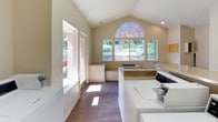 This shared laundry facility is a thoughtfully designed, single-level space that combines practicality with an airy, welcoming atmosphere. The room features striking vaulted ceilings that rise to an arched transom window, flooding the interior with natural light and creating an unexpectedly open feel for a utility space. Large rectangular windows along one wall offer views of the surrounding landscaped grounds, further enhancing the brightness of the room.

The facility is well-appointed with multiple top-loading washing machines and a bank of stacked dryers, providing ample capacity for shared use. A spacious laminate folding counter runs along the center of the room, offering generous surface area for sorting and folding laundry. The warm wood-look flooring adds a clean, modern touch throughout. Overhead fluorescent lighting ensures the space remains well-lit at all times.

Additional practical features include a bulletin board for community notices, a waste receptacle, and built-in bench seating beneath the windows. A dedicated HVAC unit keeps the room climate-controlled year-round. An exterior door provides convenient, direct access to the surrounding outdoor area. The neutral, light-toned walls and clean white appliances contribute to a tidy, functional environment that serves the needs of its users efficiently.