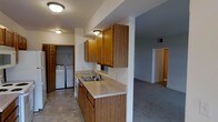 This single-floor residence is designed with practicality and comfort in mind. The kitchen serves as a central hub of the home, featuring warm oak cabinetry that extends to the upper walls for generous storage capacity. A double-basin sink sits along a continuous light-colored countertop that wraps the perimeter, providing ample workspace for meal preparation. Flush-mount ceiling lighting keeps the area well-lit throughout the day. A standout convenience of this space is the in-unit laundry area, tucked directly off the kitchen and enclosed by a full-size door — housing a side-by-side washer and dryer set on durable tile-style flooring. The kitchen opens naturally into the adjoining living area, which features neutral carpet flooring and a spacious, open layout that allows for flexible furniture arrangements. A hallway visible from the kitchen leads further into the home, connecting additional rooms and maintaining a smooth, logical flow throughout the floor plan. The neutral color palette of soft whites and grays used consistently across the space lends a clean, cohesive aesthetic. Overall, this residence offers a well-organized, move-in-ready layout that balances everyday functionality with comfortable living.