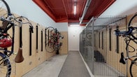 This purpose-built bicycle storage room offers a well-organized and functional space designed to meet the needs of cyclists. The room is defined by its striking red-painted exposed ceiling, which houses the building's mechanical systems and fluorescent lighting, creating a bold visual contrast against the white walls and natural plywood wall paneling.

Along one wall, a plywood-backed panel system supports multiple vertical wall-mounted bike hooks and strap-style hangers, allowing bicycles to be stored efficiently in an upright position. This maximizes floor space while keeping bikes easily accessible. Opposite this wall, a row of secured wire mesh storage cages provides additional locked storage capacity, suitable for bicycles or other equipment requiring extra security.

The flooring consists of durable concrete with a central carpet runner along the main corridor path, guiding movement through the space. Two distinct entry points are visible — a wood-paneled door with a circular porthole window and a stainless steel kick plate at one end, and a set of solid double doors with keypad access control at the other — ensuring secure and convenient entry. The space is well-lit throughout, and its practical, no-frills design prioritizes functionality, making it an ideal amenity for an active, bike-friendly community.
