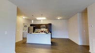This single-floor space is designed with an open-concept layout that seamlessly connects the kitchen and living area, creating a functional and inviting environment. The kitchen is a standout feature, anchored by a large peninsula with a light granite countertop that provides ample workspace and serves as a casual dining or entertaining surface. Dark espresso cabinetry lines the kitchen walls, offering generous storage, while stainless steel appliances — including a refrigerator, microwave, and range — add a modern, polished touch. Pendant lighting hangs above the peninsula, complementing the recessed and flush-mount ceiling fixtures that provide consistent illumination throughout the space. The flooring is a warm-toned wood-look laminate that extends throughout the open area, contributing to a cohesive and contemporary aesthetic. The living area benefits from the open design, offering generous room for furnishing and personalization. White walls keep the space feeling bright and neutral. A washer and dryer unit is visible in an adjacent utility area, adding practical in-unit convenience. Overall, the space combines modern finishes with a clean, functional layout.
