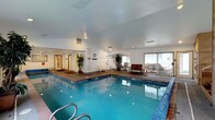 This single-level indoor pool facility centers around a generously sized swimming pool with clear, blue-tinted water illuminated by underwater lighting. The pool is framed by a tiled surround that transitions into a wood-toned deck area, providing ample space for poolside use. Entry to the water is facilitated by stainless steel handrails along the steps.

The surrounding deck is furnished with a variety of seating options, including white lounge chairs, a bench, and a small table-and-chair set, offering comfortable spaces for relaxation and socializing. Decorative potted trees are positioned throughout the space, adding a touch of greenery to the interior environment.

The room features a vaulted white ceiling with recessed lighting, contributing to a bright and open atmosphere. Large sliding glass doors and windows along one wall allow natural light to filter in and provide views of the exterior. The walls are finished in a neutral white tone, keeping the space feeling clean and airy. Safety equipment is visibly mounted on the walls, and a clock is posted near the entrance, reflecting the facility's practical, well-organized layout.
