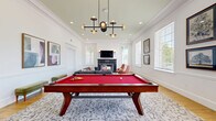 Located in Jacksonville, Florida, this well-appointed recreational lounge offers a thoughtfully designed single-floor layout that blends classic architectural charm with contemporary furnishings. The space is anchored by a striking red-felt billiards table set atop a patterned area rug over warm hardwood flooring, creating an inviting focal point for social gatherings. Soaring ceilings with detailed crown molding and recessed lighting lend an airy, refined ambiance, further elevated by a bold, sculptural multi-arm pendant chandelier. A cozy seating area at the far end of the room features a large sectional sofa, terracotta accent chairs, and a sleek dark marble fireplace topped with a wall-mounted television, making it ideal for relaxed conversation or entertainment. Abundant natural light pours in through multiple large windows dressed with blinds, offering views of lush greenery outside. The walls are adorned with paneled wainscoting and curated artwork, contributing to the space's polished aesthetic. A dramatic black arched entryway with transom detail connects to an adjoining area with high-top bar seating, adding a distinct architectural element. The overall design strikes a balance between classic Southern elegance and modern comfort.